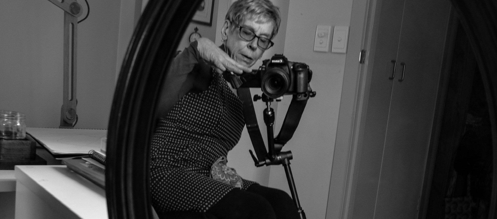 a circular mirror showing a lady leaning into view of the mirror frame with a camera propped on a tripod in front of her and crutches to her side