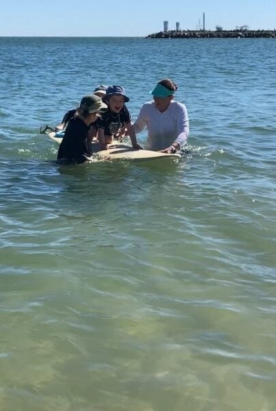A young boy atop a surfboard in shallow waters with two women standing in the water beside him, a third adult is obscured behind the board. They are surrounded by water with a rocky break water in the far distance. 