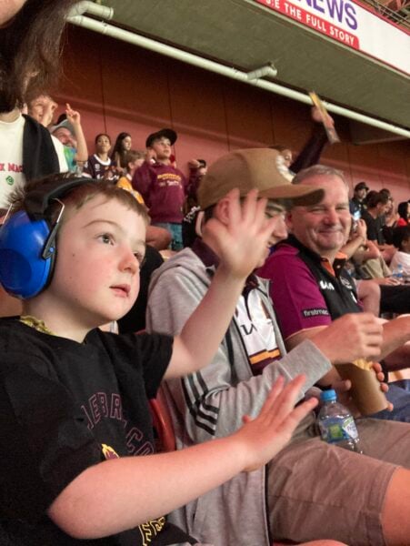 Within the maroon spectator stands filled with people in maroon jerseys, a young boy wearing headphones in the foreground with his hands waving sits next to his family. 