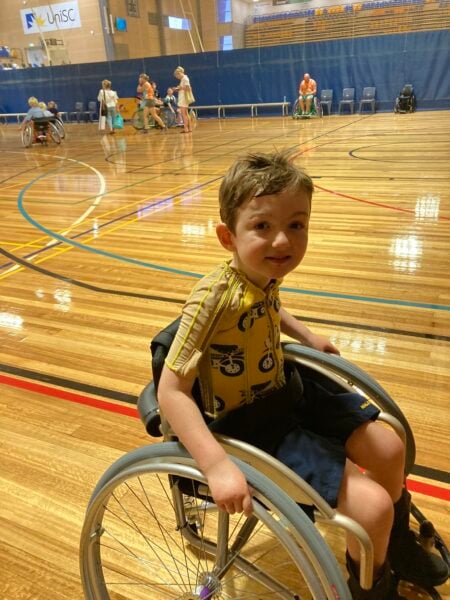 A young boy using a wheelchair on a basketball court with coloured lines snaking behind him. There are around 5-6 other wheelchair basketballers behind him on the far side of the court. 