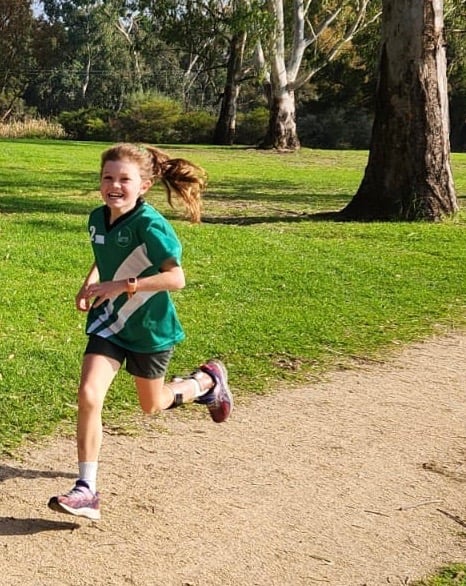 A young girl in a green shirt with white stripes, brace on her left leg, which trails behind her runs on a gravel path with a big smile next to green grass and trees.