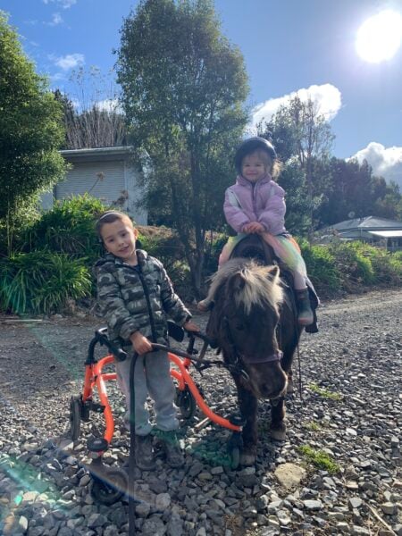 A smiling young boy wearing a camo jacket and using a walking frame is leading a pony. A girl toddler wearing a rainbow tutu sits proudly on a saddle on the pony's back.