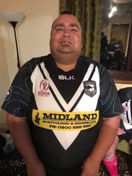 A man stands proudly puffing his chest with arms by his side. He is wearing a black jersey with a large white "V" design with logos "Fesitval of World Cups" on his right chest, "Physical Disability Rugby League New Zealand" on the left chest and sponsor logo in the centre.