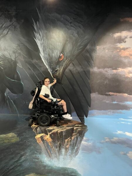 A teenager sits in a power wheelchair, smiling and looking toward someone off camera. Their chair is positioned on a giant mural covering the entire floor and wall, making it look like they are perched on a floating rock. Their hand is touching the beak of an enormous black raptor bird.