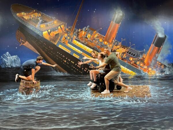 A teenager in a power wheelchair playfully enacts a scene from "Titanic" with their parents and sibling. The ship is sinking in the background on a giant mural that covers the entire floor and wall. The family appears to be perched on wooden objects floating in the ocean.
