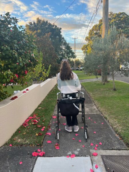 A teenager uses a walker to stroll down a suburban footpath. The sun is setting and there are trees lining the path and red flowers have fallen on the ground. 