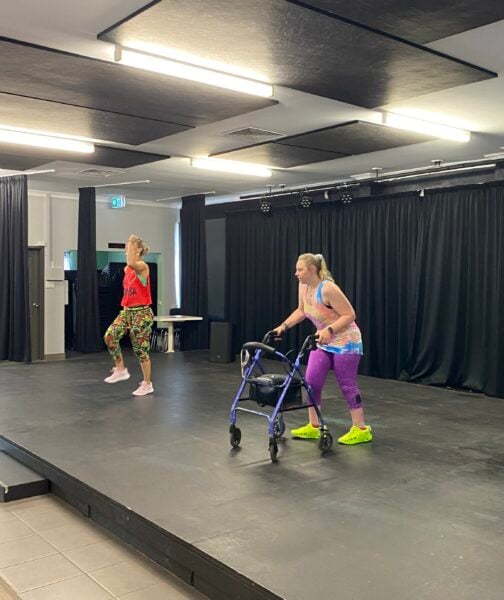 A woman with brightly coloured fitness clothes and using a four wheeled walker stands on stage. She is looking towards an instructor who is demonstrating Zumba moves.