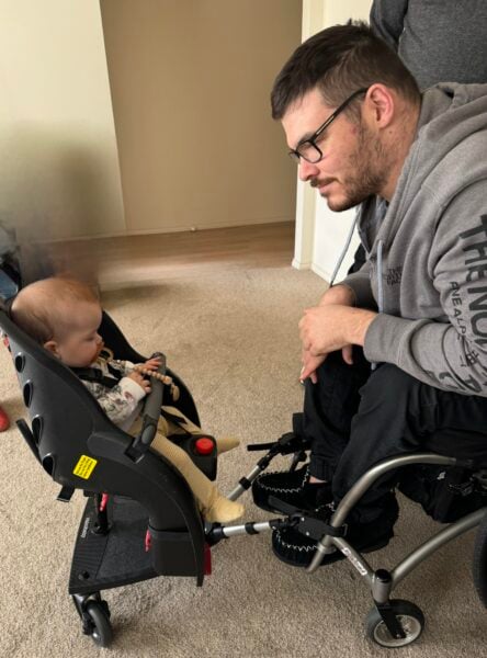 A man in a grey jacket sitting in a wheelchair looking at a baby sitting in a bicycle seat attached to his wheelchair. 