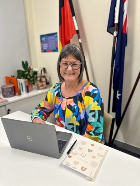 Frances in a bright colourful top sits at a desk with a grey laptop. Aboriginal and TSI flags behind her
