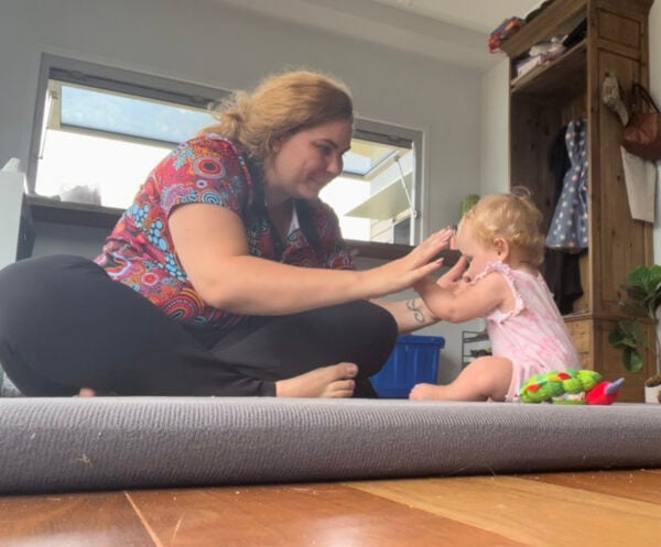 Martika sits cross legged on a mat reaching her hands towards a sitting baby with some toys around them