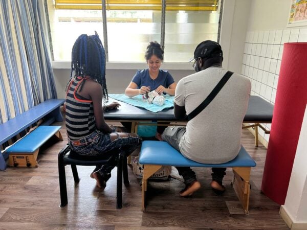 Two parents observe a baby being treated by a clinician. Baby is on a soft table, parents seated with backs to viewers, clinician on the opposite side facing camera. Windows to outside building behind her.