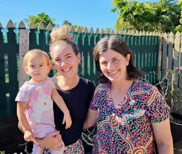 Two adults and a toddler, who is being held, stand together smiling in front of a green fence and blue sky above them. 