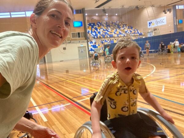 A selfie photo of a woman and a child in a wheelchair on a basketball court with lots of colourful ground lines.