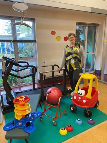 A lady stands with a hand on a kid's treadmill in a therapy gym with a car, ball, walker and other toys in front of her.