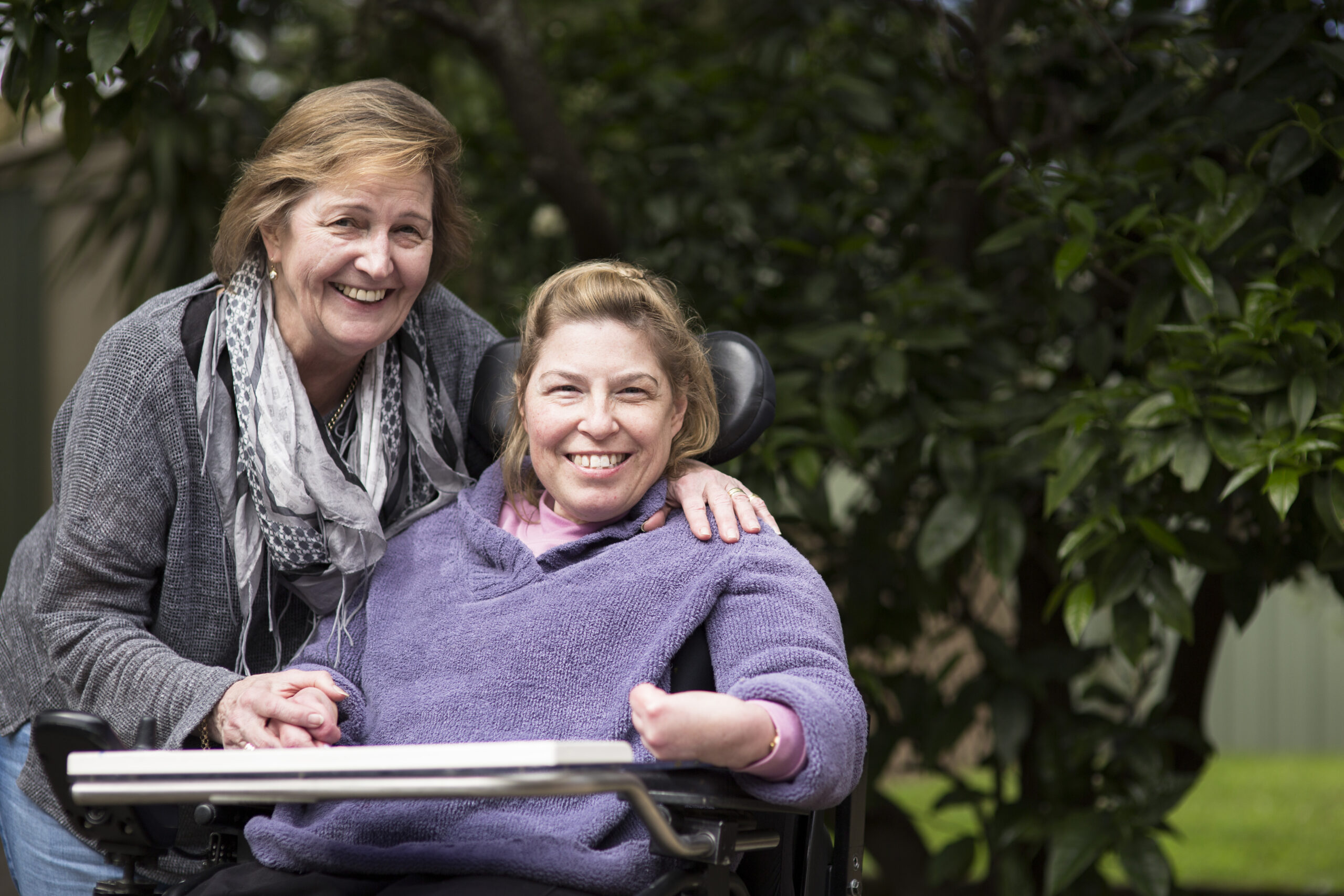 This image shows two women   smiling, located in a sunny area underneath some trees. One woman extends her arm to rest it on the shoulder of the other woman.